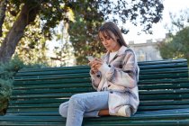 Modern millennial female in stylish spring outfit sitting on bench and browsing on mobile phone while resting on urban street looking away — Stock Photo
