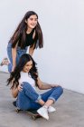 Fröhlicher Teenager mit gekreuzten Beinen fährt Skateboard mit zufriedenen weiblichen Geschwistern tagsüber auf Gehweg — Stockfoto
