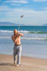 Полная длина взрослого спортивного мужчины, смотрящего в сторону и делающего вокруг тела передачу с весовой пластинкой на ленте, согреваясь на солнечном побережье — стоковое фото