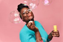 Щаслива афроамериканка з закритими очима в синьому одязі і розмахуючи мильними бульбашками на рожевому тлі в студії — стокове фото