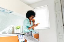 Desde abajo joven mujer afroamericana en ropa casual comer manzana y navegar por Internet en el teléfono móvil mientras está de pie en la cocina casera - foto de stock