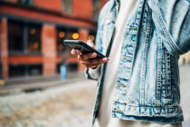 From below crop anonymous African American guy in denim trendy jacket surfing modern mobile phone during city stroll — Stock Photo