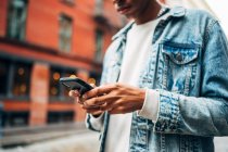 From below crop anonymous African American guy in denim trendy jacket surfing modern mobile phone during city stroll — Stock Photo