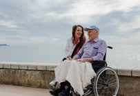 Довольна взрослая дочь и пожилой отец в инвалидной коляске охлаждения на набережной против моря вместе летом — стоковое фото