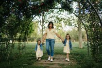 Corpo cheio de jovem segurando as mãos de filhas adoráveis em roupas semelhantes enquanto caminham juntas no gramado verde no parque de verão — Fotografia de Stock