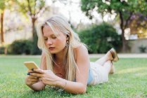 Unemotional charming female lying on grass in park and listening to music in headphones in summer — Stock Photo