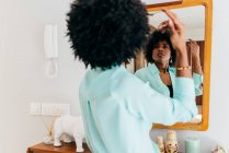 На задньому плані - вродлива молода афроамериканка в повсякденному одязі, що торкається кучерявого волосся, стоячи в кімнаті і дивлячись на дзеркало. — стокове фото