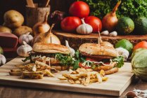 Hamburgers appétissants aux légumes et escalopes placés sur une planche de bois avec frites dans la cuisine — Photo de stock