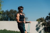 Сильный спортсмен в спортивной одежде и солнцезащитных очках с эластичной лентой на газоне при солнечном свете — стоковое фото