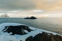 Сніжна скеля, що виходить на острів посеред бурхливого моря з хмарним небом у зимовий день на Лофотенських островах (Норвегія). — стокове фото