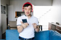 Deleitado adolescente latino en auriculares en videollamada en el teléfono móvil mientras está de pie cerca del sofá en casa y mirando a la cámara - foto de stock