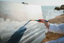 Crop young woman standing on grassy coast near sand and ocean in sunny day while drawing picture with brush on canvas on easel — Stock Photo