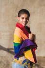 Joven bisexual étnica femenina con bandera multicolor mirando a la cámara y representando símbolos LGBTQ en un día soleado - foto de stock