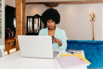 Jeune pigiste afro-américaine en tenue décontractée buvant du café assis à table et travaillant sur un projet avec ordinateur portable à la maison — Photo de stock