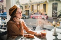 Französin in Baskenmütze sitzt am Tisch im Café mit aromatischem Glas Kaffee und frisch gebackenem Croissant — Stockfoto