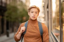 Confident ethnic male model with blond hair and in trendy turtleneck standing in city and looking at camera — Stock Photo