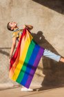 Vue latérale de la jeune femme bisexuelle ethnique avec jambe levée et drapeau multicolore debout sur la passerelle sur fond beige — Photo de stock