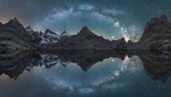 Breathtaking night scenery of rough rocky mountains with snow near calm lake with smooth water surface reflecting sky with shiny Milky Way — Stock Photo