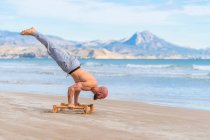Side view full length of male athlete performing handstand with parallel bars on sandy coast with ocean waves on background — Stock Photo