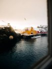 Vue imprenable sur les cabines jaunes et rouges situées sur le littoral enneigé contre le ciel gris avec des oiseaux depuis la fenêtre du navire sur les îles Lofoten, Norvège — Photo de stock