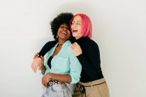 Cheerful young pink haired woman embracing positive African American girlfriend in stylish outfit while having fun together on white background — Stock Photo