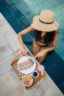 From above of anonymous female tourist in straw hat sitting in pool while cutting delicious crepe with chocolate sauce — Stock Photo