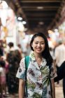 Happy Asian female traveler in tropical shirt with backpack smiling at camera while standing on bazaar against blurred crowd in Doha — Stock Photo