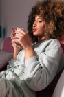 Femme afro-américaine avec les cheveux bouclés assis sur le canapé et boire une boisson chaude à la maison — Photo de stock