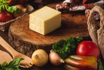 Bloco de queijo em suporte de madeira perto de cebolas cruas e saco de tecelagem contra espátulas orgânicas e folhas de manjericão — Fotografia de Stock