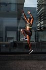 Seitenansicht der starken schwarzen Frau springen hoch in der Nähe Glaswand des modernen Gebäudes während der Übung auf der Straße der Stadt an sonnigen Tag — Stockfoto