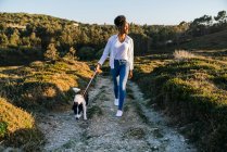 Corpo inteiro de mulher étnica feliz com Border Collie cão andando juntos na trilha entre colinas gramadas na noite de primavera ensolarada — Fotografia de Stock