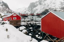 Camino de madera que va cerca de la pared de la choza en la aldea costera cerca de la cresta de montaña nevada en el día de invierno en las Islas Lofoten, Noruega - foto de stock