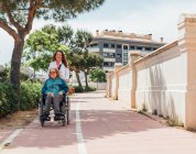 Glückliche erwachsene Frau blickt in die Kamera, während sie im Sommer mit ihrer Mutter im Rollstuhl auf der Straße in der Stadt spaziert — Stockfoto