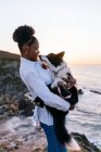 Vista lateral de la propietaria afroamericana sosteniendo lindo perro Border Collie feliz mientras disfrutan del tiempo juntos cerca del mar ondeando al atardecer - foto de stock
