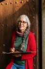 Artista donna anziana deliziata con tavolozza di colori e pennelli in piedi vicino alla porta di legno e guardando la fotocamera — Foto stock