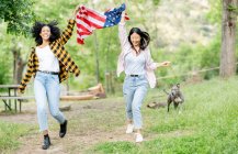 Alegre lesbiana multirracial pareja de hembras corriendo con nacional americano bandera a lo largo de camino en bosque y sonriendo mirando cámara - foto de stock