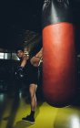 Joven centrado hombre asiático entrenamiento kick boxing realizar golpes patadas mientras se ejercita con saco de boxeo pesado en un gimnasio moderno - foto de stock