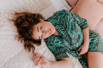 Высокий угол спокойной женщины в домашней одежде, отдыхающей на мягкой кровати дома и смотрящей в камеру — стоковое фото