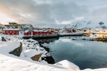 Холодное море со спокойной водой, расположенное рядом с прибрежным поселением и снежным горным хребтом в пасмурный зимний день на Лофских островах, Норвегия — стоковое фото