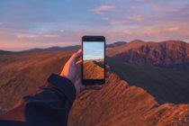 Caminante masculino anónimo de cultivos tomando fotos de las tierras altas en el teléfono inteligente al atardecer en Gales - foto de stock