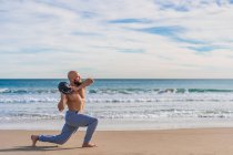 Vue latérale de l'homme sportif sans chemise portant kettlebell et faisant des fentes de marche sur la côte sablonneuse avec des vagues de l'océan sur le fond — Photo de stock