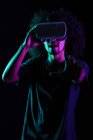Homem latino animado com penteado afro e em óculos VR experimentando realidade virtual em fundo preto em estúdio com luzes de néon — Fotografia de Stock