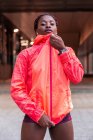 Ethnisch athletische junge Frau blickt in die Kamera und reißt rote Jacke auf der Straße — Stockfoto