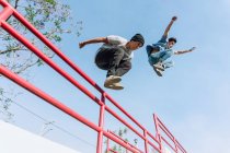 Desde abajo intrépidos amigos masculinos saltando por encima de la barandilla de metal en la ciudad mientras realizan acrobacias de parkour en un día soleado - foto de stock