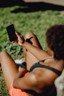 Irreconocible mujer afroamericana en ropa deportiva sosteniendo teléfono inteligente mientras está sentado en la hierba en un día soleado - foto de stock