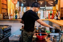 Vista laterale dell'uomo in bandana in piedi al bancone e il ramen di cottura nel moderno caffè asiatico — Foto stock