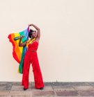Femme afro-américaine joyeuse dans des vêtements élégants avec bouteille de boisson alcoolisée et drapeau coloré regardant loin sur fond clair — Photo de stock