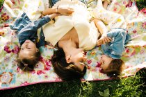 Top view of happy young woman and adorable little sisters in similar dresses lying on blanket on green grass while spending summer day together in park — Stock Photo