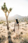 Comprimento total de fotógrafa feminina com câmera em pé na terra do deserto do parque nacional contra a árvore Joshua verde na Califórnia EUA — Fotografia de Stock