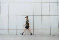 Vista laterale della donna allegra in abbigliamento casual e scarpe da ginnastica che cammina guardando la fotocamera con le braccia tese sopra la passerella piastrellata in città — Foto stock